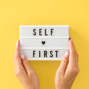 Hands holding a sign that says, Self First.
