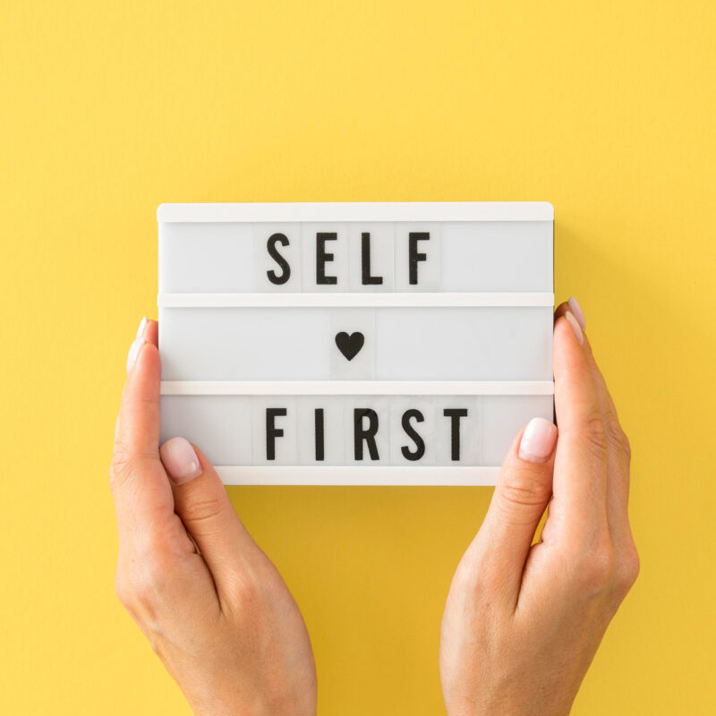 Hands holding a sign that says, Self First.